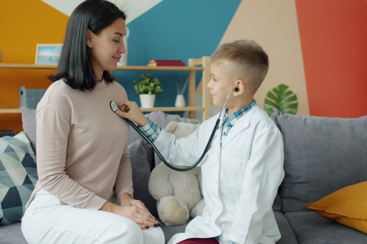 Little boy in white gown is playing with mom examining her with stethoscope while woman is smiling and breathing enjoying game with cute child.
